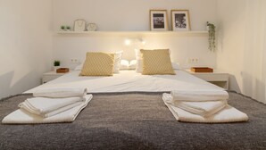 Ein ordentlich bezogenes Bett mit weißen Leinen und zwei Kissen, ein Nachttisch mit einer Pflanze und ein Wandregal mit gerahmten Fotos und kleinen Dekorationsgegenständen.