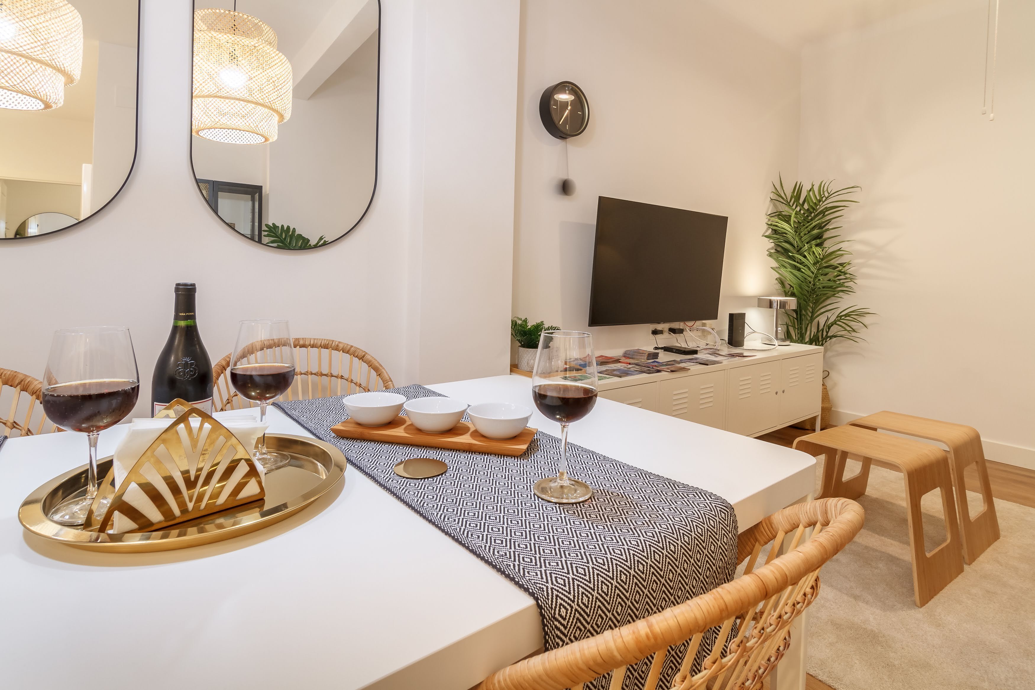 Een eethoek met een witte tafel, rieten stoelen, een dienblad met glazen en een fles, en een televisie op de achtergrond.