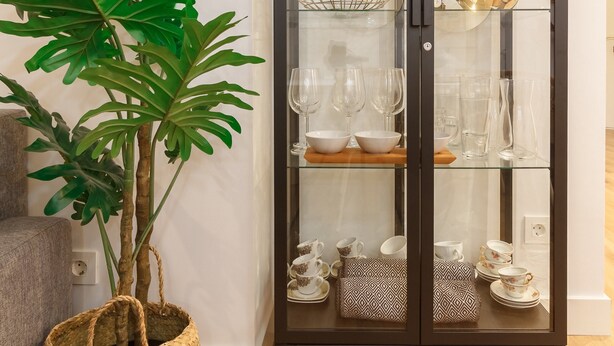 Ein Zimmer mit einer gläsernen Vitrine, die Glaswaren und Dekorationsobjekte zeigt, einer großen Topfpflanze und einem gewebten Korb am Boden.