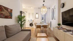 Ein modernes Wohnzimmer mit einem grauen Sofa, einem Holztisch und einem Essbereich mit einem weißen Tisch und Korbstühlen. Ein großer Flachbildfernseher ist an der Wand montiert, und im Hintergrund ist eine Küche zu sehen.