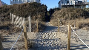 Een zandpad langs het strand leidt naar een houten hek en een gebouw met een witte gevel en meerdere ramen.