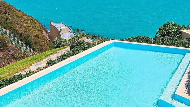 A rectangular swimming pool with clear blue water, surrounded by a landscaped area with greenery and a view of the ocean. - Stunning Resort Apartment at Las Casitas (Fajardo)