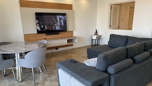 A modern living room with a grey sofa, a TV mounted on a wooden panel, a dining table with chairs, and a small side table. - Stunning Resort Apartment at Las Casitas (Fajardo)