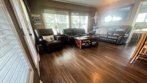 A living room with a wooden floor, leather sofas, a coffee table, and a window with blinds. - Fisherman’s Getaway with Sunset Views & Room for 11 (Jewett)