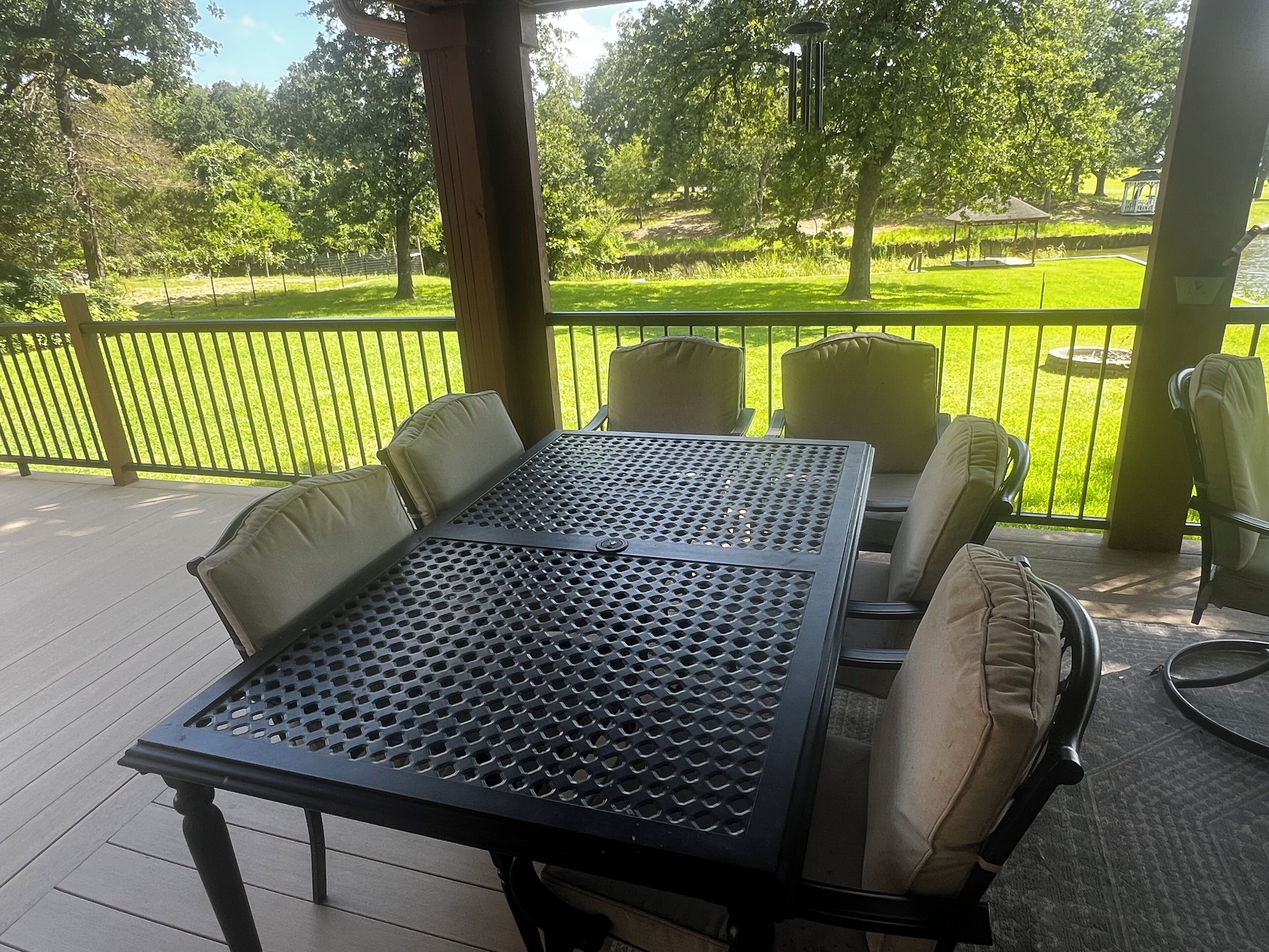 Um alpendre coberto com uma mesa e cadeiras de metal, vista para uma área gramada com árvores e um gazebo ao fundo.