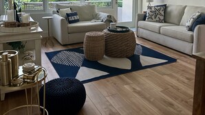 En moderne stue med sofa, sofabord og skammel, et stort vindue med udsigt til bygninger og træer, samt et dekorativt spejl på væggen.