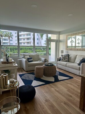 A modern living room with a sofa, coffee table, and ottoman, a large window with a view of buildings and trees, and a decorative mirror on the wall.