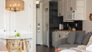 Una cocina moderna con armarios blancos, un microondas y una encimera con un bol de limones. Hay una zona de comedor con una mesa de vidrio y sillas, y una sala de estar con un sofá gris y una cama blanca.