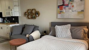 Un dormitorio moderno con una cama grande, un sofá gris, un reposapiés redondo pequeño y una zona de cocina con armarios y un microondas.