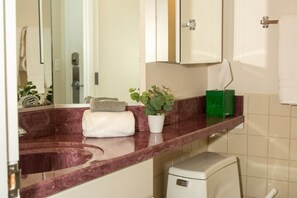 Une salle de bain avec un comptoir en marbre, des toilettes blanches, un distributeur de savon vert et une plante en pot.