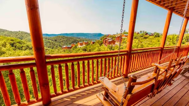 A wooden deck with a swing chair, a railing, and a view of green hills and buildings in the distance. - NEW LISTING-IN-Cabin Pool-Arcades-King Beds (Sevierville)