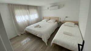 Siisti, moderni hotellihuone, jossa on kaksi erillistä sänkyä, suuri ikkuna läpikuultavilla verhoilla, seinälle asennettu ilmastointilaite ja yöpöytä lampulla.