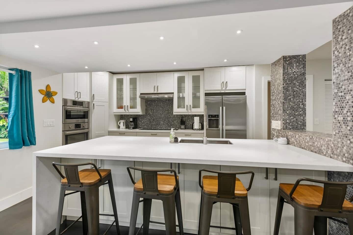 Une cuisine moderne dotée d’un grand îlot central, de tabourets de bar, d’appareils électroménagers en acier inoxydable et d’un revêtement mural décoratif composé de mosaïques.