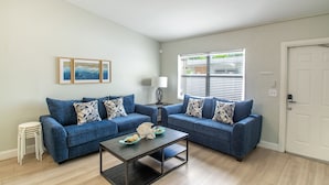A living room with two blue sofas, a coffee table, and a side table with a lamp. There are framed pictures on the wall, a window with blinds, and a white door. - Beautiful remodeled condo. Steps away from area's top nightlife/1 mi. from beach (Delray Beach)