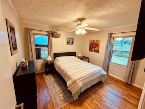 A bedroom with a bed, a nightstand, a lamp, a ceiling fan, a window, and a painting on the wall.