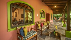 Uma área de estar coberta ao ar livre, com bancos de madeira, janelas com moldura verde e uma rede, além de uma pequena mesa com uma planta.