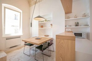 Una cocina con una mesa de madera, sillas, una lámpara colgante, estantes con vajilla y un microondas.