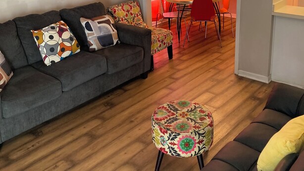 Ein Wohnzimmer mit grauem Sofa, bunten Kissen, Holzboden und Deckenventilator. Eine Essecke mit roten Stühlen und Tisch ist ebenfalls zu sehen.