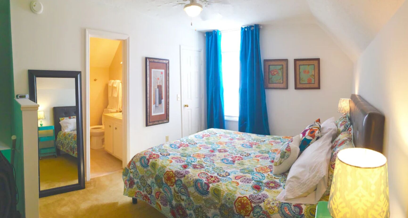 A bedroom with a bed, a bedside lamp, a mirror, and a door leading to a bathroom.