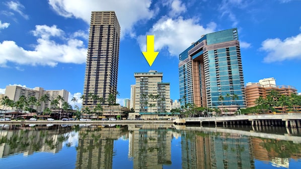 Um edifício alto refletido na água, cercado por palmeiras e outros edifícios.