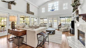 A spacious living room with a fireplace, large windows, and a high ceiling.