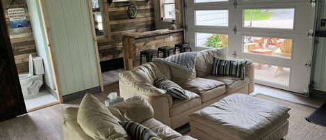 A cozy living area with a beige leather sofa, a wooden bar area, and large windows providing a view of the outdoors.