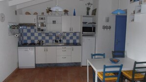 Une cuisine compacte avec des meubles blancs, un crédence carrelée, un four à micro-ondes et un évier. On y trouve une table à manger avec des chaises, et une porte bleue en arrière-plan.