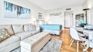 A modern, well-lit room with a sofa, ottoman, and a bed with a blue headboard. There is a dining area with a table and chairs. - Waterfront Condo w/Pool Access (Fort Walton Beach)