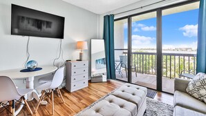 A modern living room with a flat-screen TV, a dining area with a table and chairs, a sofa, and a balcony with a view of the sky and palm trees. - Waterfront Condo w/Pool Access (Fort Walton Beach)