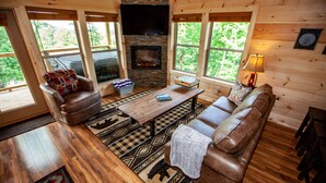 Interior acogedor de una cabaña con chimenea, muebles de madera y ventanas amplias que ofrecen vistas al exterior.