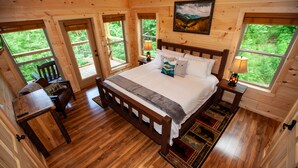 Un dormitorio de cabaña de madera con una cama grande, dos mesitas de noche con lámparas, una silla y vistas al exterior a través de amplios ventanales.