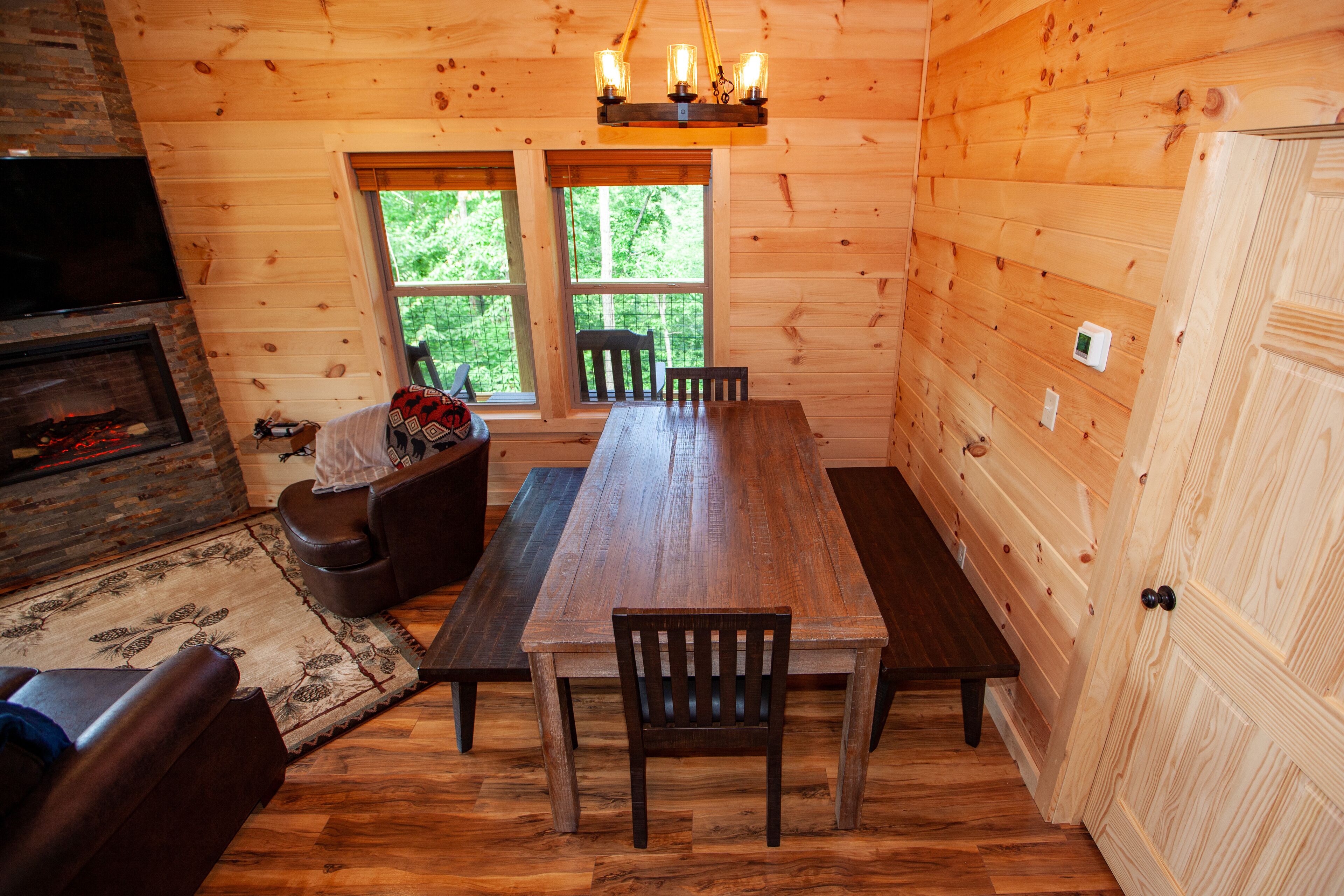 A wooden interior with a dining table, chairs, a fireplace, and a large window.