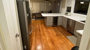 Una cocina con suelo de madera, armarios blancos, un refrigerador, un lavavajillas y una ventana con persianas.