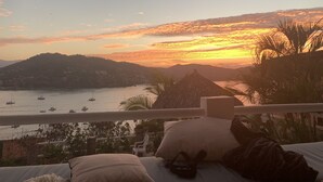 En balkon med udsigt over en solnedgang over vand, både og et bakket landskab.