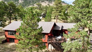 Une cabane à deux étages avec une terrasse, entourée de pins et située au pied d’une chaîne de montagnes.