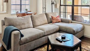 A beige sectional sofa with a throw blanket, a small wooden coffee table, and a window with blinds. - 2-Bedroom Condo in the Foothills, La Encantada, Galleries and Restaurants! (Tucson)