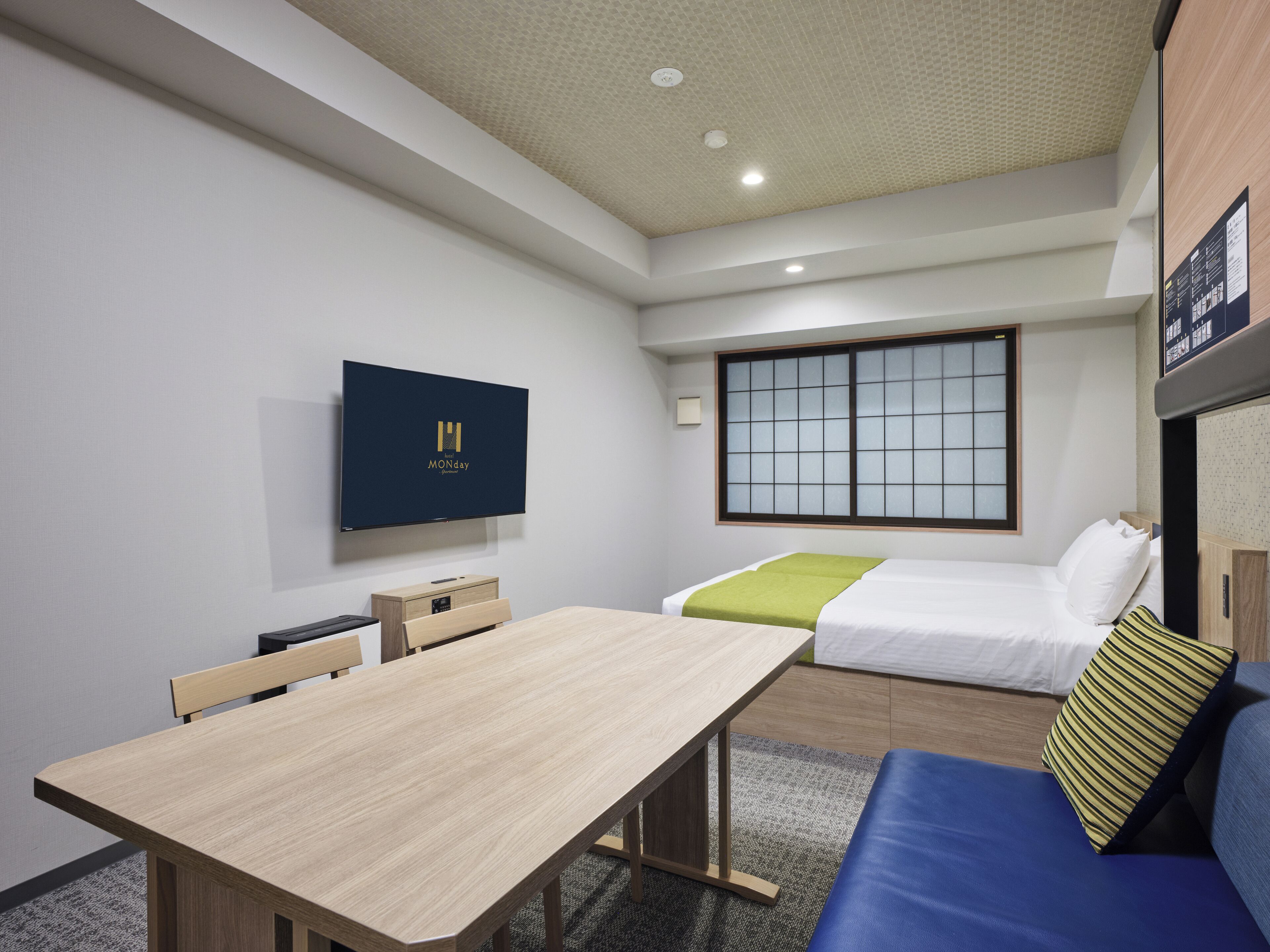 A hotel room with a wooden table, a bed with a green blanket, a blue sofa, a TV mounted on the wall, and a window with a grid pattern.