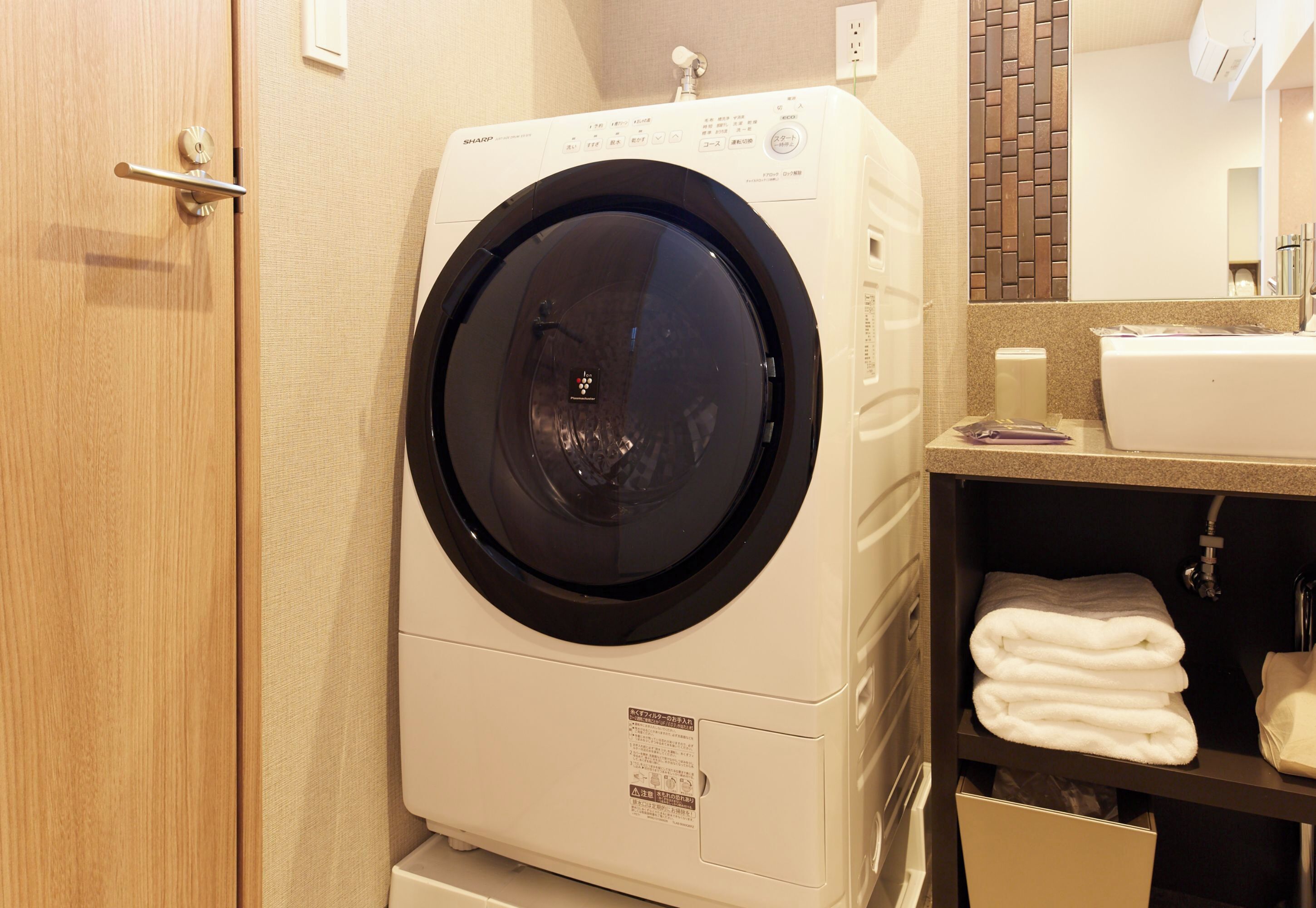 Une machine à laver compacte et moderne dans une chambre d’hôtel, avec une serviette soigneusement pliée posée sur le comptoir et un miroir au-dessus du lavabo.