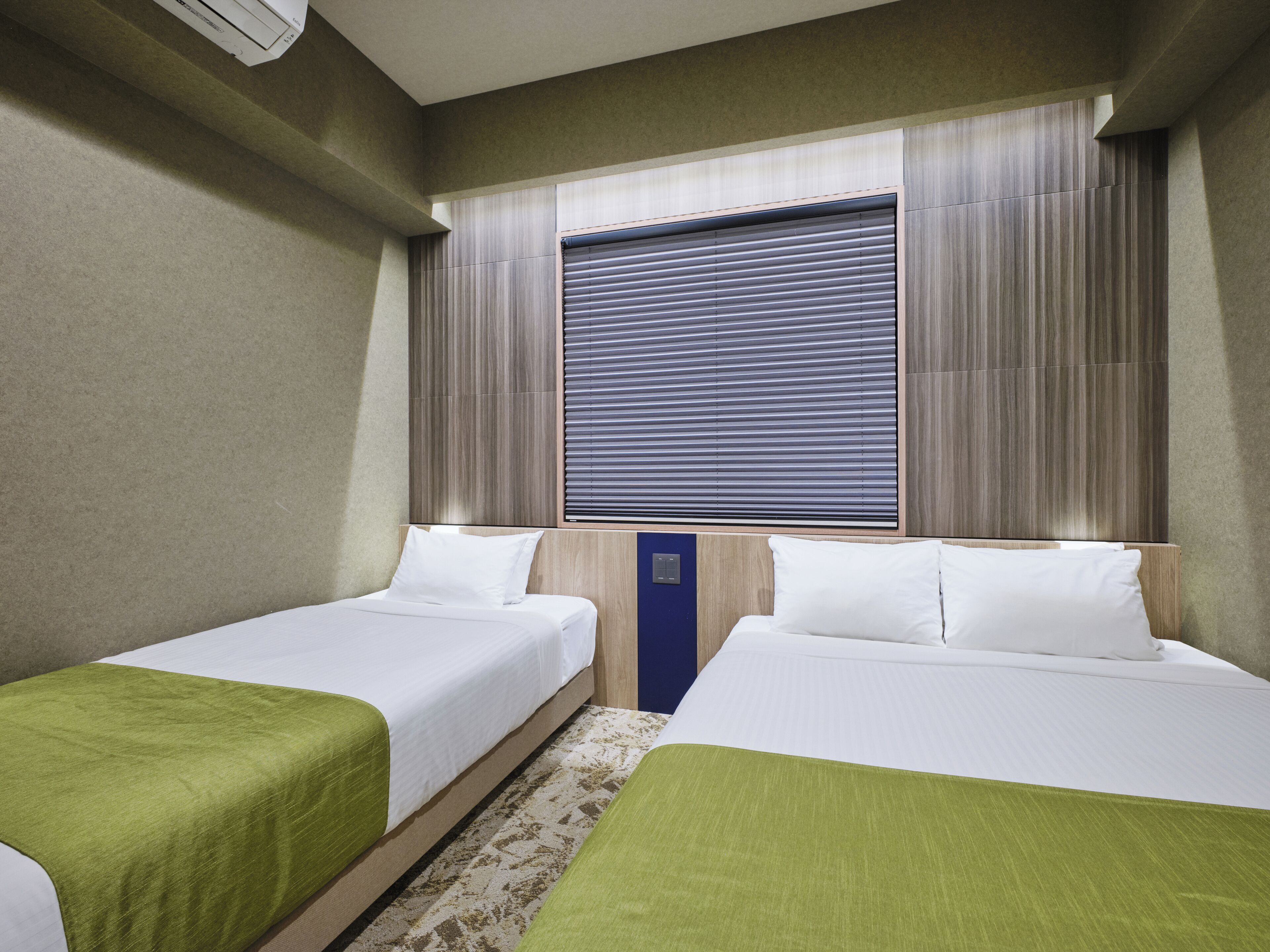 Ein Hotelzimmer mit zwei Betten, einem großen Fenster mit Jalousien, einer Wand-Klimaanlage und Teppichboden.