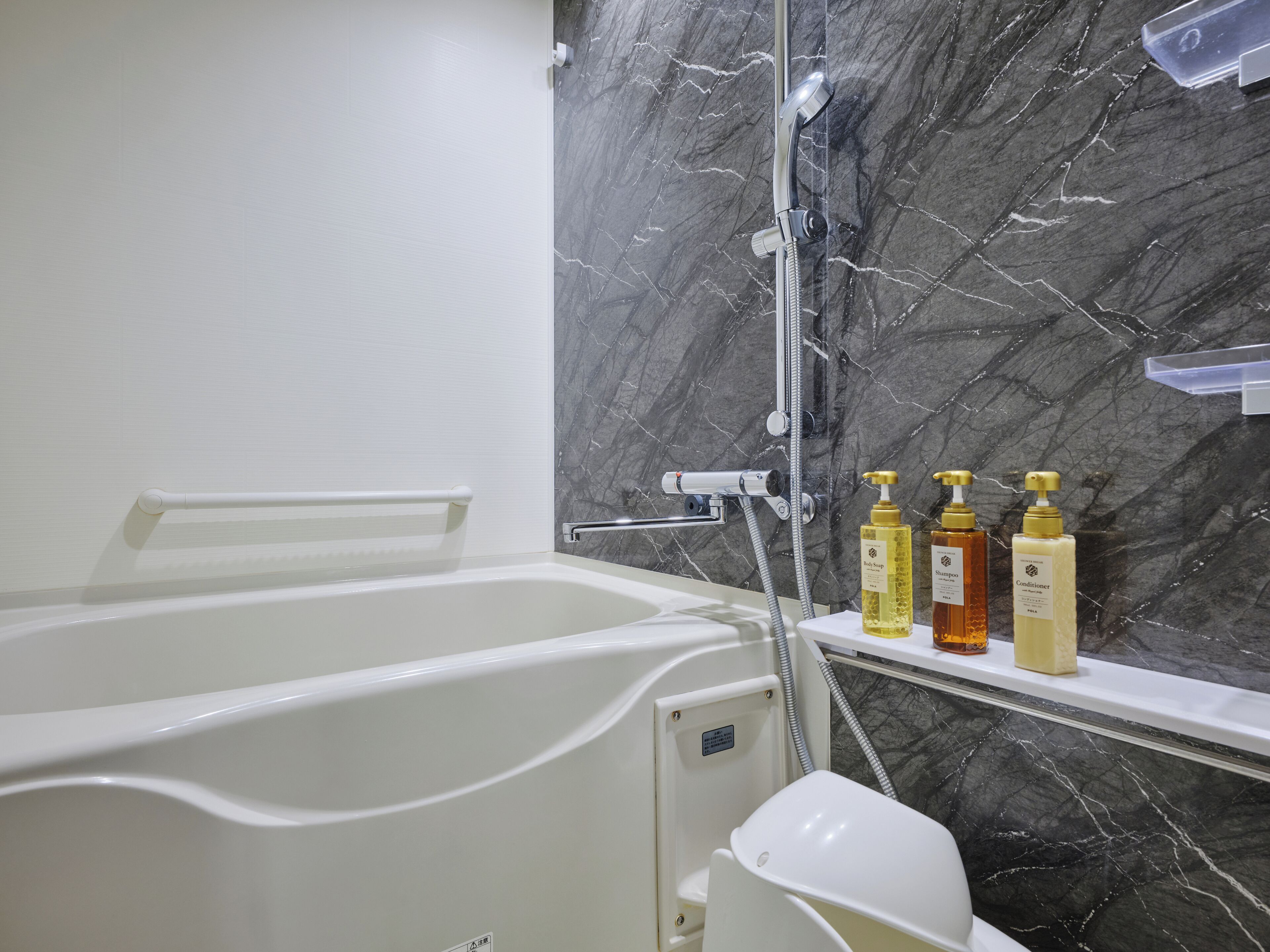 A modern bathroom with a bathtub, a shower, and a marble wall. There are three bottles of toiletries on a shelf.