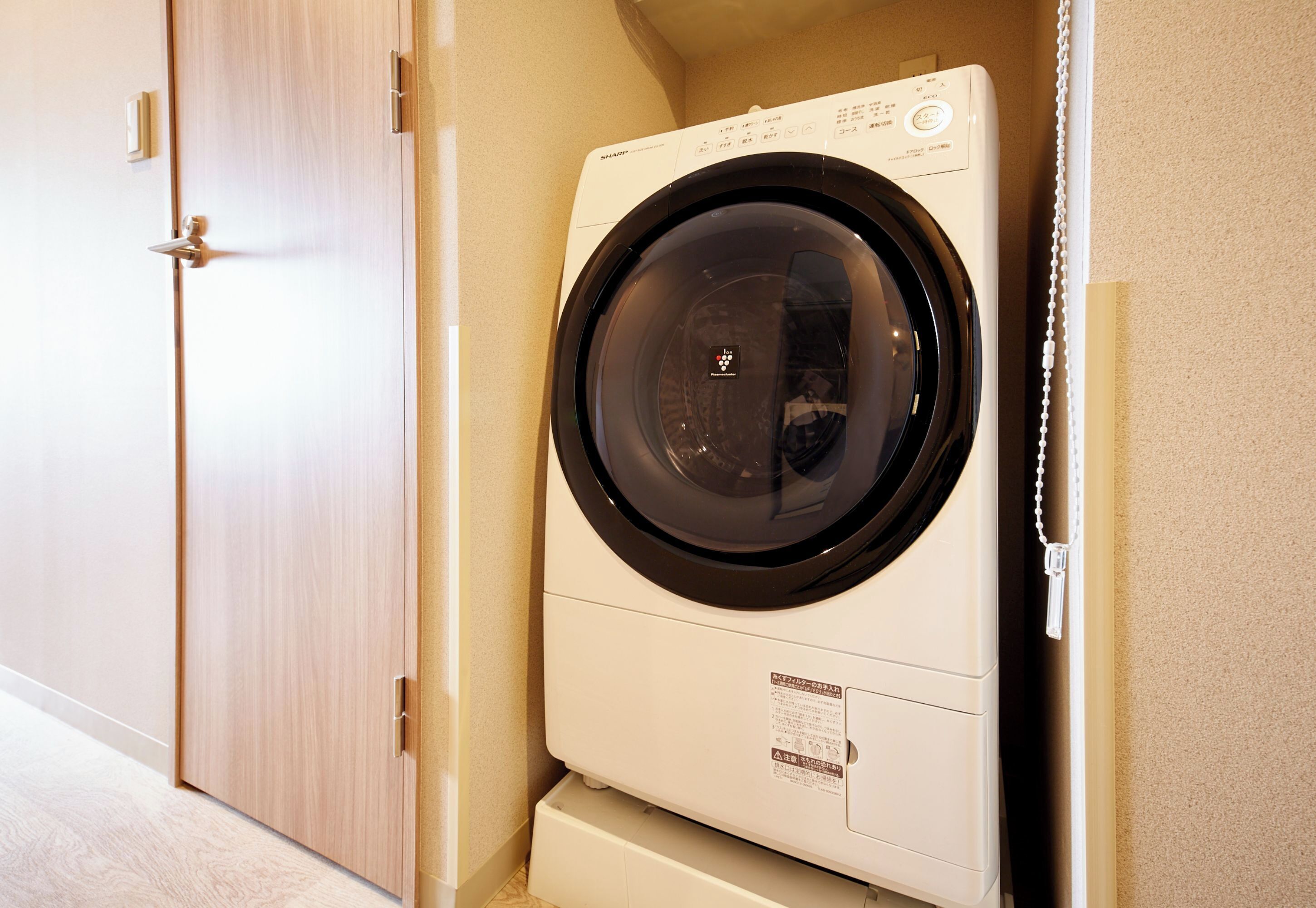 A compact, white front-loading washing machine in a small laundry room with a wooden door and beige walls.