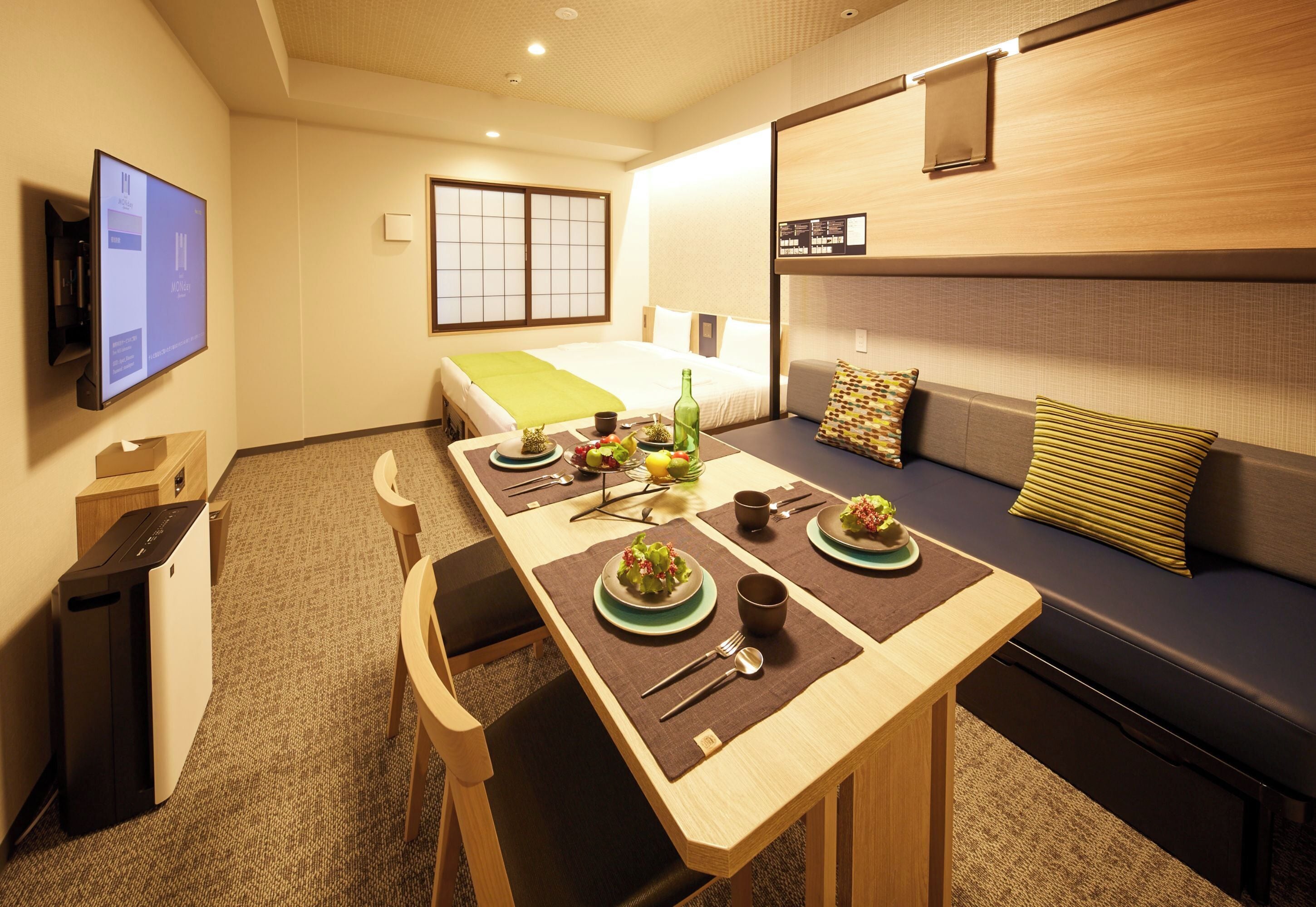 A hotel room with a dining table set for two, a sofa, a TV, and a bed with a green blanket.