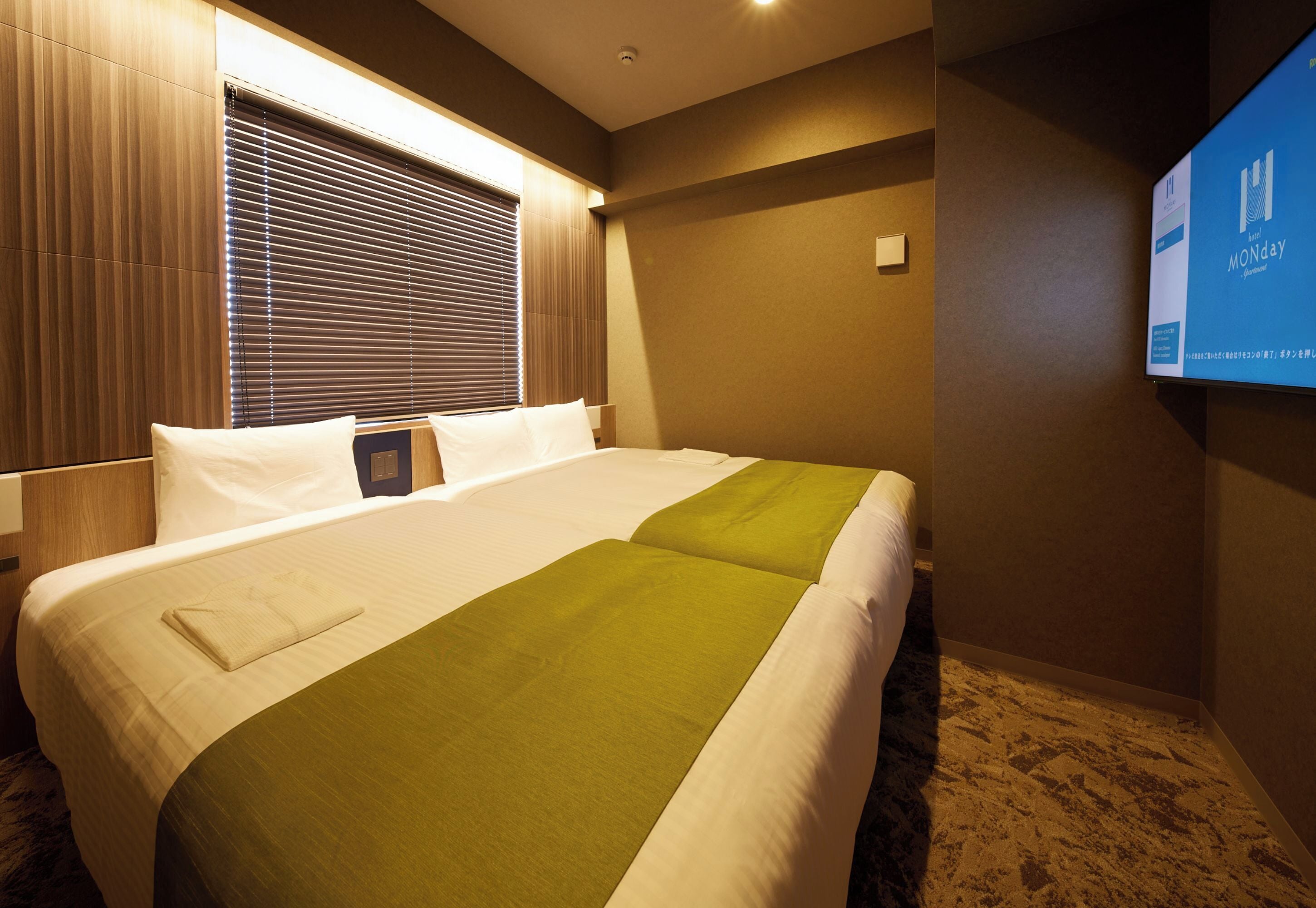 A hotel room with a large bed, a television on the wall, and a window with blinds.