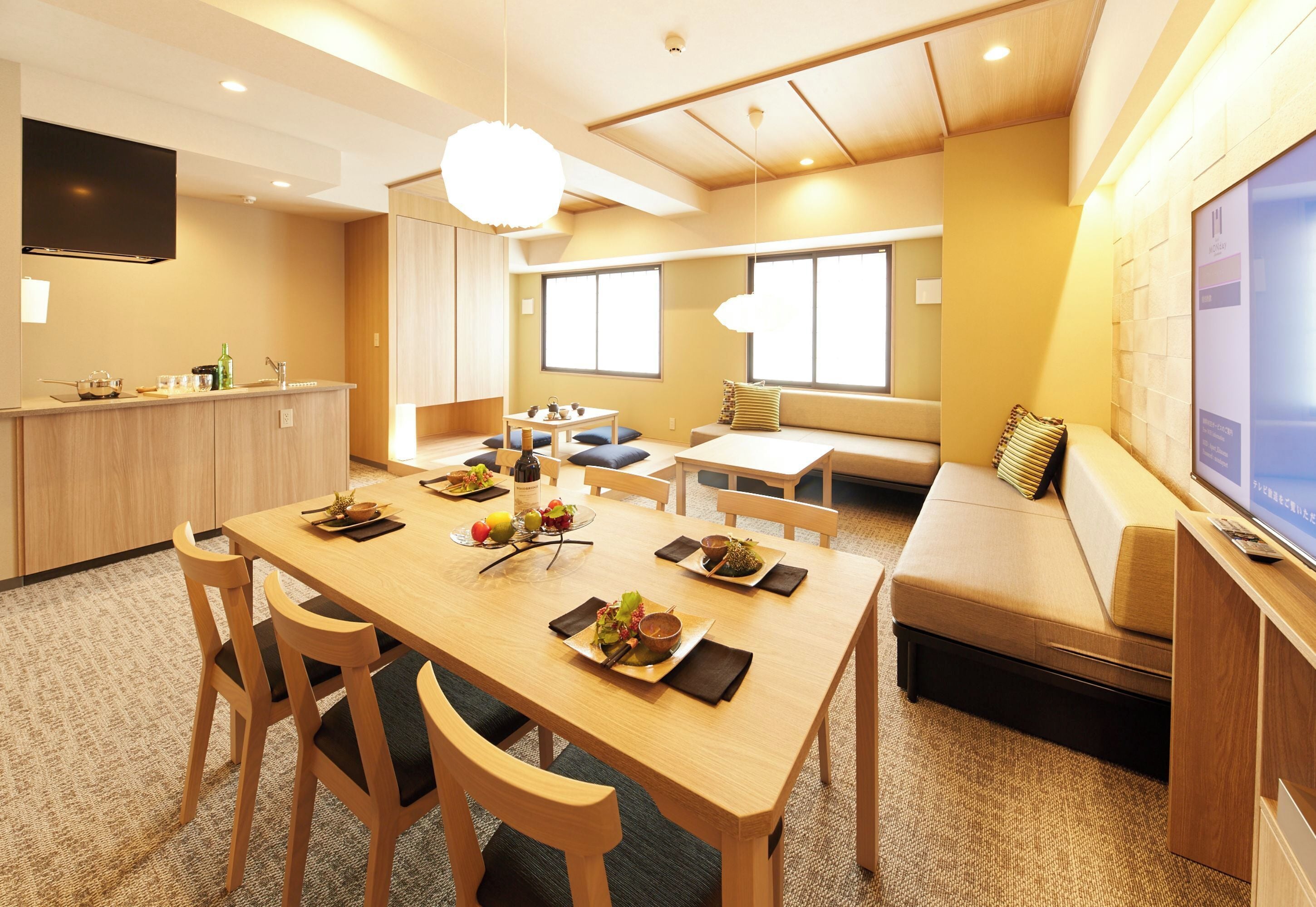 A modern hotel room with a wooden dining table, chairs, a sofa, and a TV.