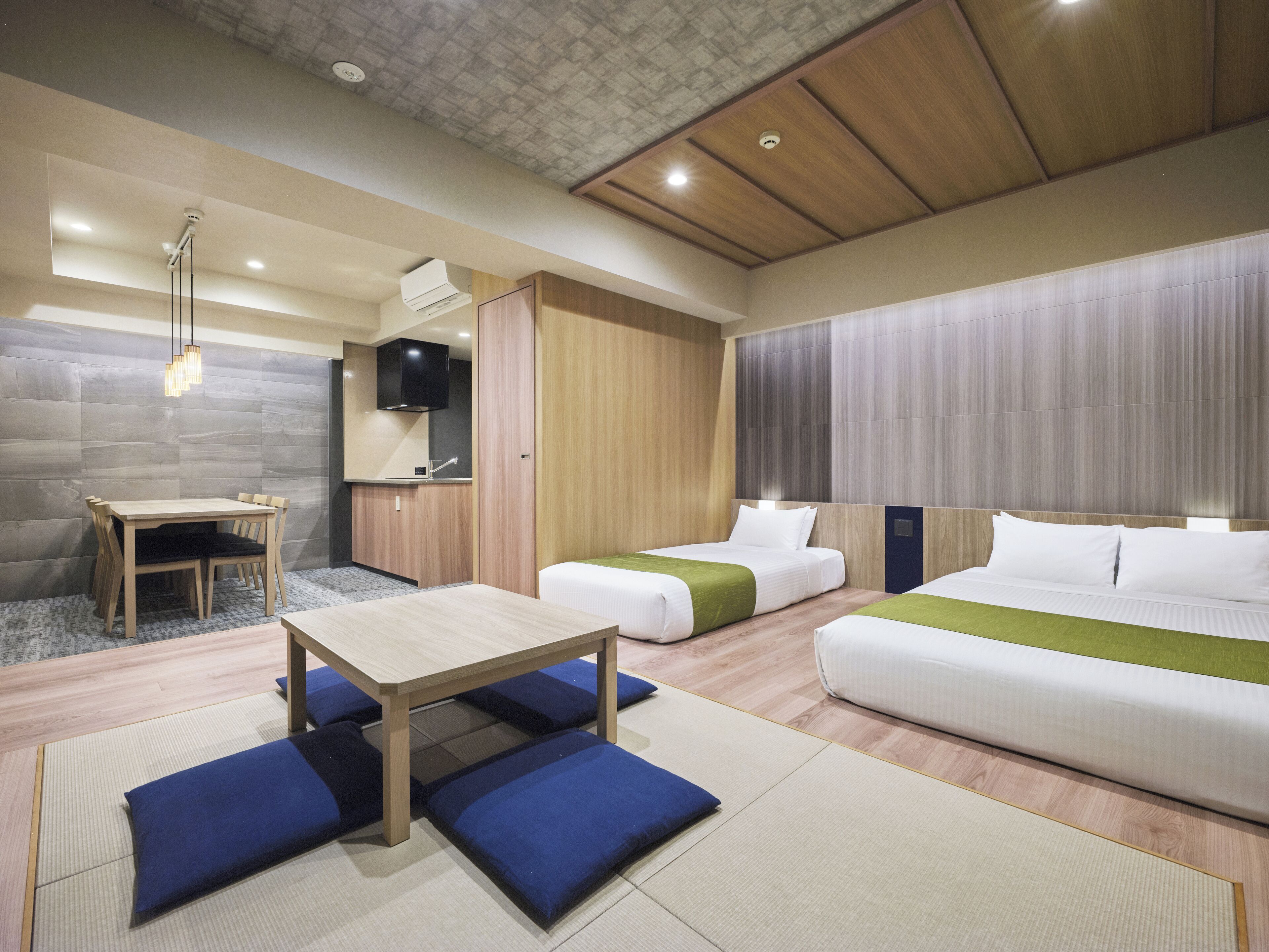 A modern hotel room with a wooden ceiling, a dining area with a table and chairs, two beds with green and white bedding, and a TV mounted on the wall.