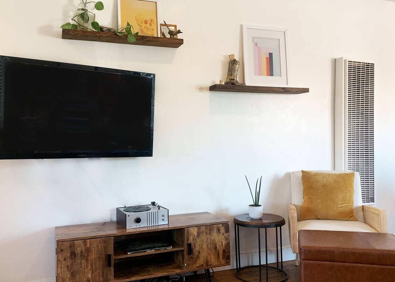 Un saló amb televisió de pantalla plana, moble de roba de fusta, taula petita rodona amb una planta, sofà amb coixí i prestatgeria de paret amb objectes decoratius.