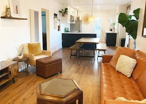 A modern living room with a sofa, ottoman, and coffee table, a dining area with a table and chairs, and a kitchen in the background. - Beautiful BOHO Home by Beach, Downtown + Shops (San Diego)
