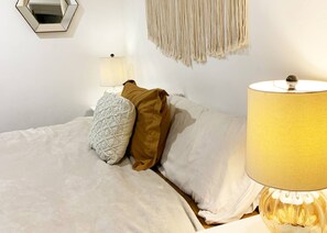 A bed with white bedding, a patterned pillow, and a brown pillow. There is a wall-mounted lamp with a yellow shade and a decorative wall hanging. - Beautiful BOHO Home by Beach, Downtown + Shops (San Diego)