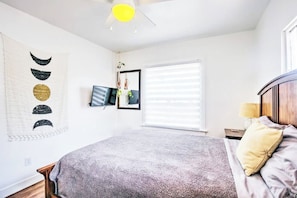 A bedroom with a bed, a bedside table, a lamp, a window with blinds, a wall-mounted TV, and a ceiling fan. - Beautiful BOHO Home by Beach, Downtown + Shops (San Diego)
