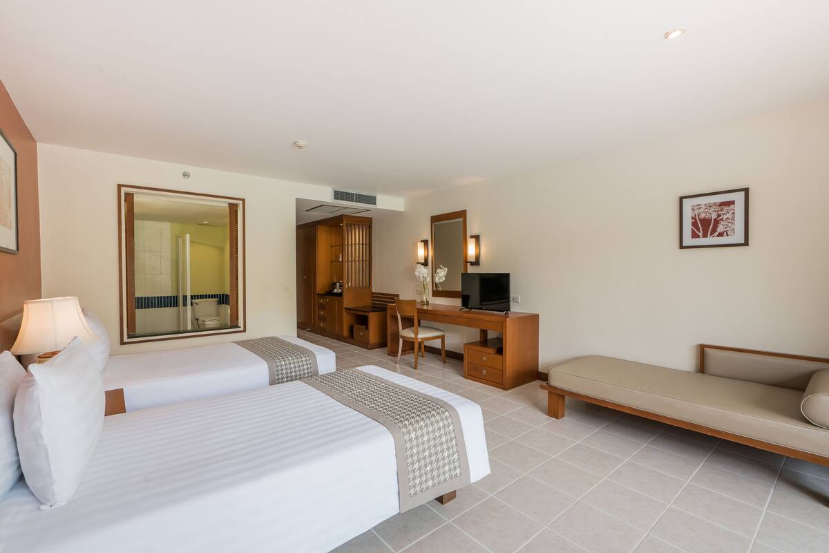 Ein Hotelzimmer mit zwei Betten, einem Schreibtisch, einem Sessel, einer Couch, einem Fernseher und einem durch eine offene Tür einsehbaren Badezimmer.